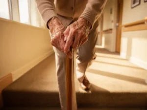 Oudere handen grijpen houten wandelstok terwijl ze voorzichtig trappen afdalen in zorgcentrum met zachte middagzon door ramen
