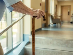 Oudere handen grijpen houten wandelstok vast terwijl ze voorzichtig stenen trappen navigeren in moderne zorgfaciliteit gang
